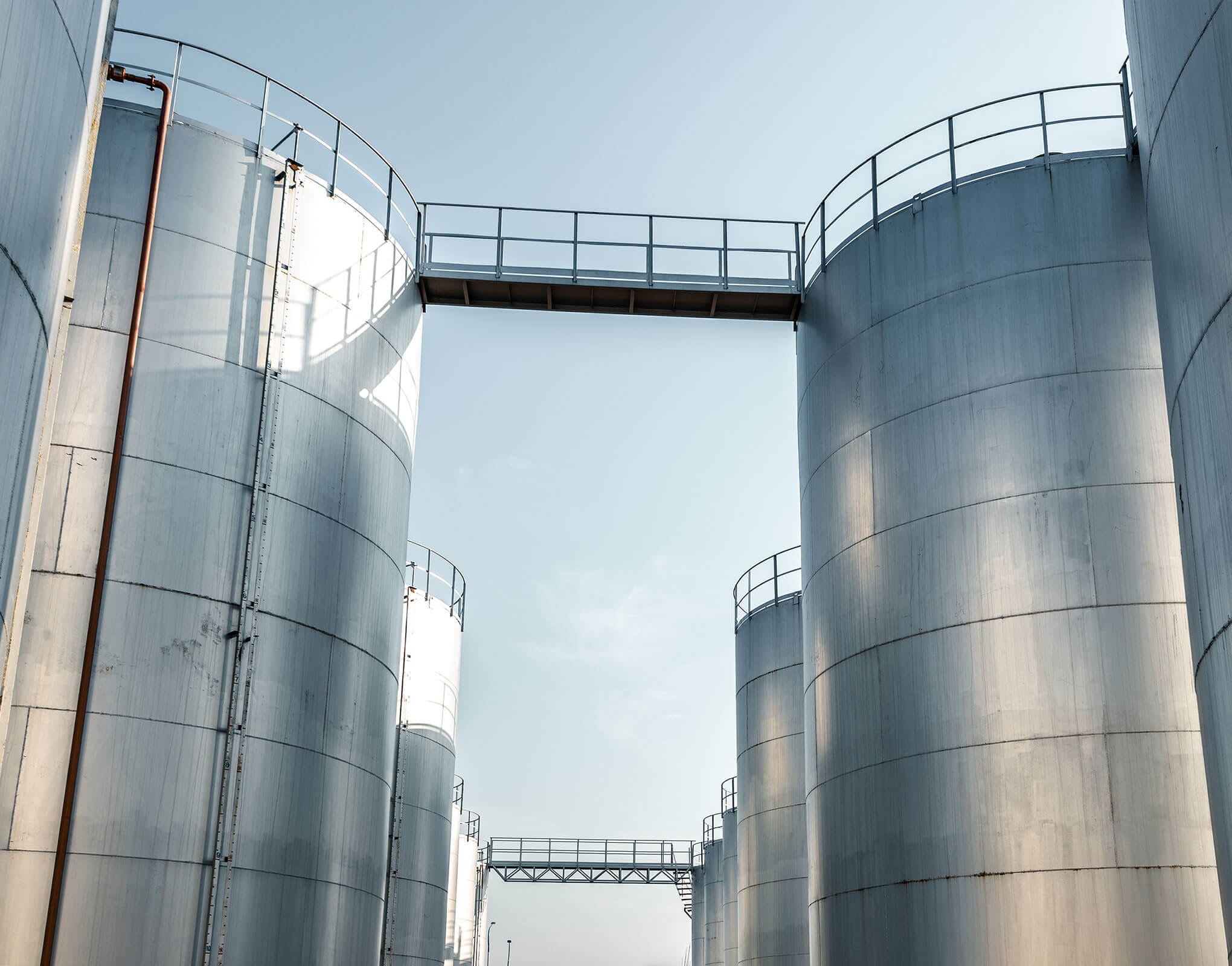 View of large silver industrial storage tanks with a walkway between them against a clear sky