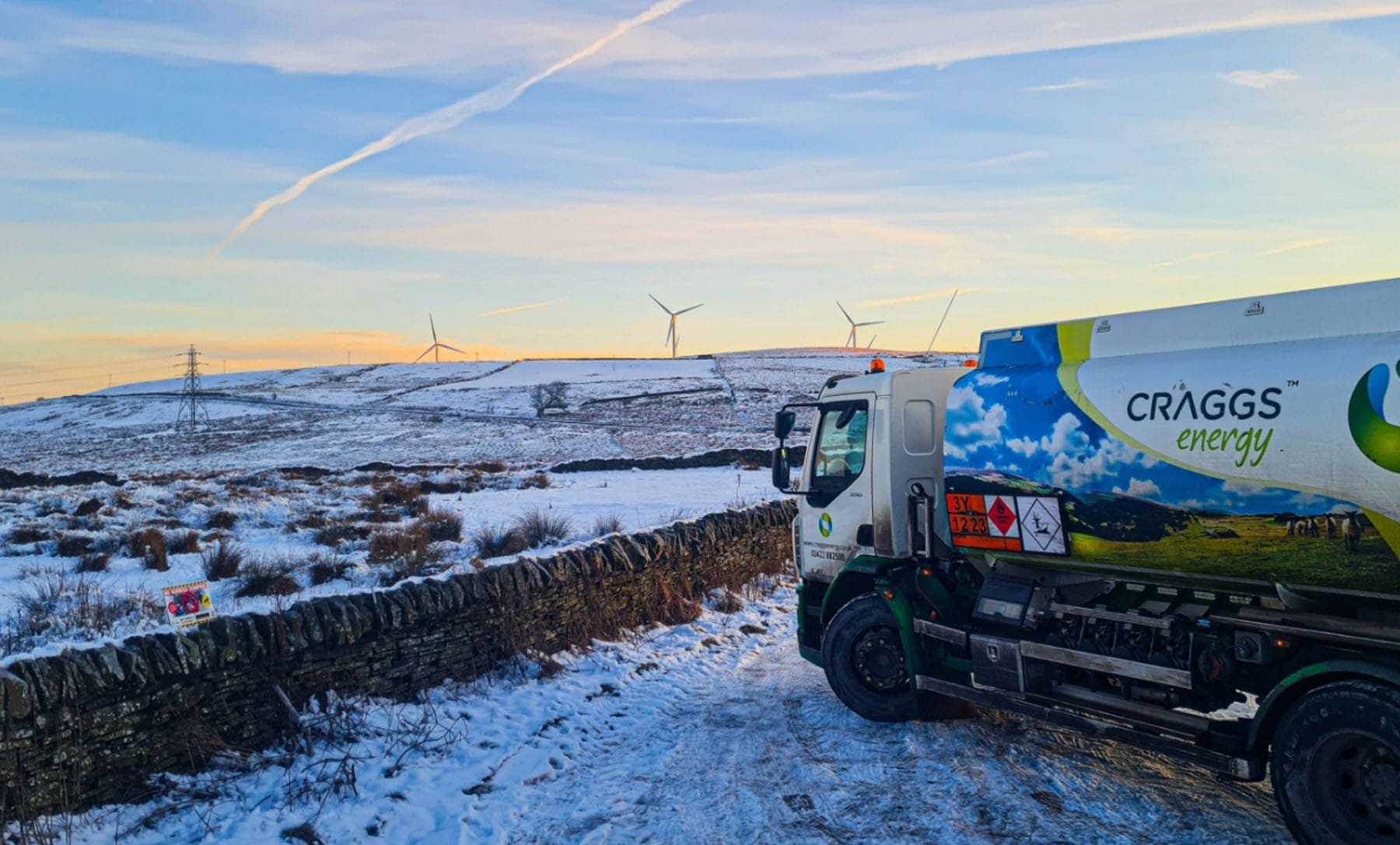 Cragg's Energy Truck on Snowy Landscape with Wind Turbines A Cragg's Energy truck parked on a snowy hillside, with wind turbines visible in the background during sunset.